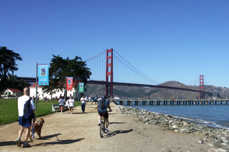 People walking and biking on flat dirt trail next to San Francisco Bay with Golden Gate Bridge in background